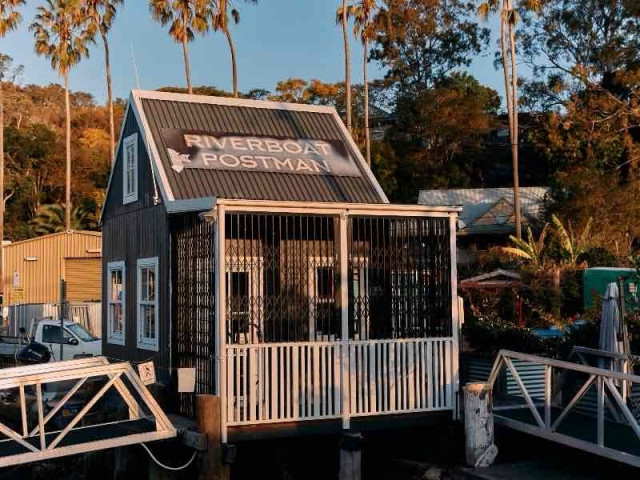 A small building with a sign reading "Riverboat Postman" on the roof, situated at the water's edge with surrounding trees and a dock.