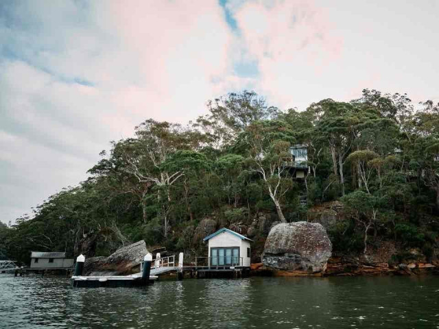 A small waterfront house with a dock sits on a rocky, tree-covered shoreline under a cloudy sky.