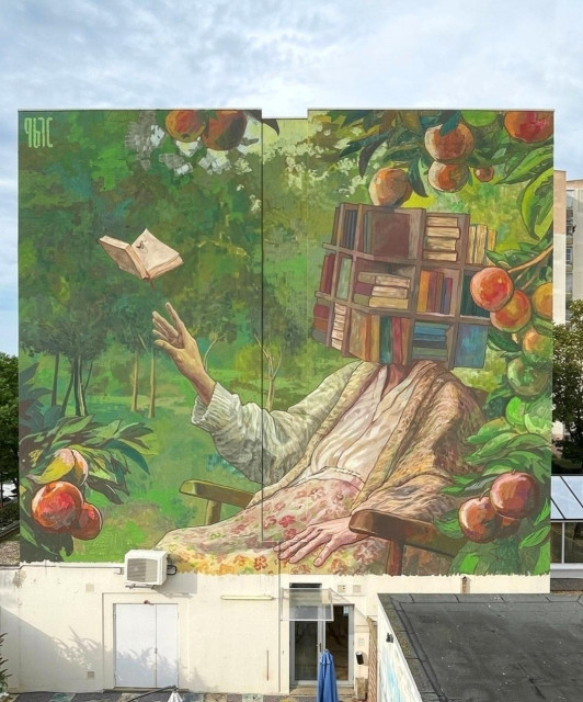 Streetartwall. A fantastic mural of a woman reading has been painted on the outside wall of a four-storey modern building. The main color is green with some bold colors. The mural shows a woman in a skirt, blouse and vest in pastel colors who has a square shelf cube full of books instead of a head. With one hand, she reaches for a book, which flies away directly in front of her like a bird. The female figure with the book head is surrounded by apple trees, nature and its green colors.
Info: The artist imagined an old woman sitting in her garden, trying to find memories that are slowly disappearing. Memories of an imaginary world, like those found in books.