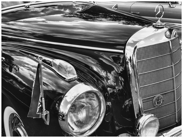 Das Schwarzweissfoto zeigt eine Schräg- oder Seitenansicht der Frontpartie eines historischen Automobils der Marke Mercedes aus dem Jahr 1956. Am rechten Bildrand thront das Mercedes Emblem auf dem Chrom-Kühler, in der  linken Bildecke ein Kotflügel mitStander und Flagge. Der Blick folgt einer Diagonalen über die schwarzglänzende Motorhaube zum Mercedes-Stern in der oberen rechten Bildecke.