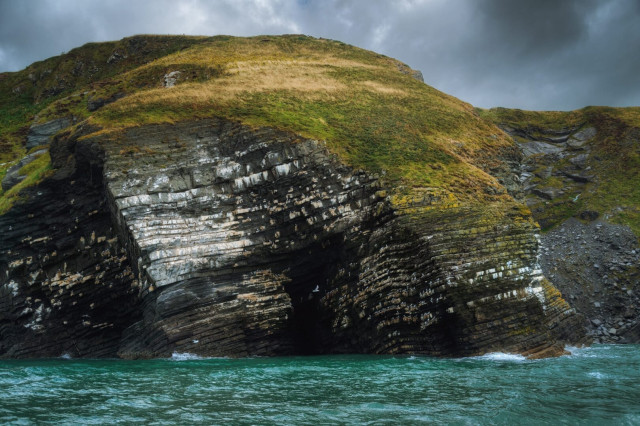 A dramatic seascape showcasing the rugged cliffs of New Quay Head, as viewed from a boat. The towering cliffside dominates the frame, revealing striking layers of sedimentary rock formations in shades of grey and brown. The cliff face is deeply furrowed and weathered, with grass-covered slopes atop the rocky outcrop. Turquoise waters of the Irish Sea lap at the base of the cliff, their choppy surface hinting at the area's dynamic coastal environment. Overhead, a moody sky with grey clouds adds to the atmospheric and wild beauty of this Welsh coastline.