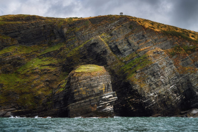 A striking view of the rugged coastline near New Quay, showcasing the dramatic geology of the Welsh coast. Towering cliffs dominate the image, their layered rock formations clearly visible, telling a story of millions of years of geological processes. The cliffs are a patchwork of dark grey stone interspersed with patches of vibrant green vegetation. A distinctive dome-shaped rock formation protrudes at the base, its rounded top covered in grass. Atop the cliff, barely visible against the overcast sky, sits a small structure, likely a lookout point or lighthouse. The churning teal waters of Cardigan Bay lap at the base of these imposing cliffs, emphasising the raw beauty of this coastal landscape.