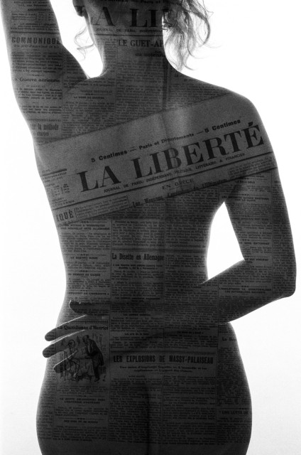 A woman's naked back from mid-buttocks to neck. Her right arm is wrapped around her back and her left is above her haid (hand off image). Her skin is superimposed with newspaper writing (in French) from the double exposure, including the title of the newspaper in block letters: LA LIBERTÉ. Black and white. 