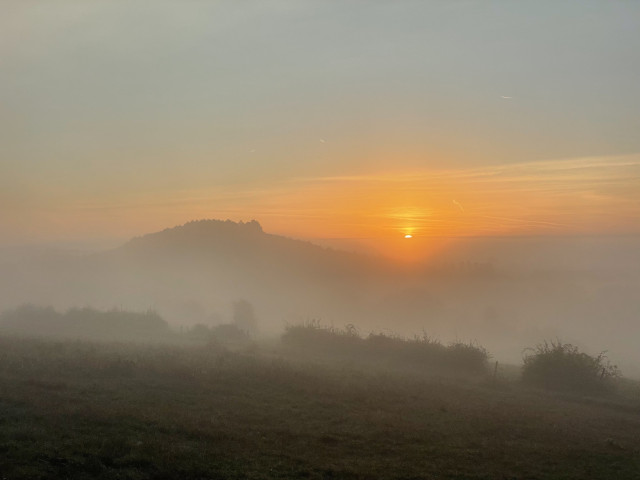 A serene landscape featuring a sunrise behind a fog-covered hill. The sky displays soft orange and yellow hues as the sun rises, casting a warm glow over the misty foreground. 
Un paysage serein avec un lever de soleil derrière une colline couverte de brouillard. Le ciel présente des teintes orange et jaune douce alors que le soleil se lève, projetant une lueur chaude sur le premier plan brumeux.