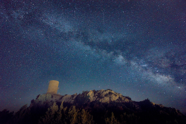 Fotografía nocturna donde en la parte inferior se ve un montículo con una pequeña atalaya en la parte izquierda y en la superior el cielo nocturno con la Vía Láctea cruzando la imagen.