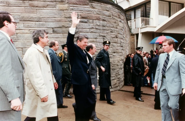 A man wears a black suit and waves his hand in the air, surrounded by other people wearing formal clothing in a vintage looking colored photo. 