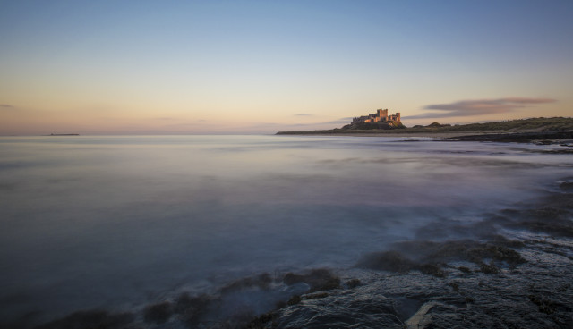 A minimalistic photo of a castle in the late evening sun. The castle is next to the sea which has a mist like appearance  caused by the use of a long exposure. 