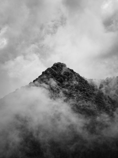 Schwarz-weiß Foto eines Berggipfels der aus dem Nebel ragt, der Himmel ist bewölkt.