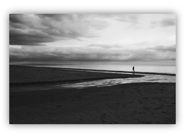 Black and white photo of a solitary figure walking along a narrow strip of land or pier that extends into the calm sea. The sky is filled with dramatic clouds, creating a reflective and moody atmosphere. The beach in the foreground is dark and textured.