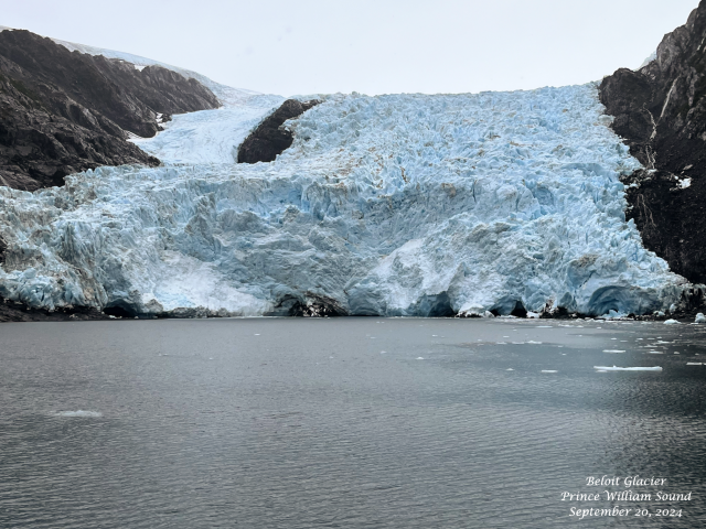 View of the face of Beloit Glacier from a boat on September 20, 2024. Deep blue ice but with imbedded rock. Rock walls on either side of glacier with cloudy skies above. Ice recently calved from the glacier is visible in the ocean in the foreground.