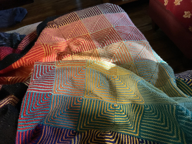 The Hue Shift Afghan, consisting of 100 mitred 2-colour blocks in all sorts of different colours, with a thick black border. Its bunched up a bit, and a ray of sunshine is shining on it.