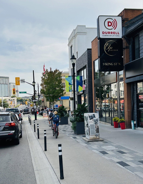 A busy urban street scene featuring cyclists on a bike lane running alongside the sidewalk.