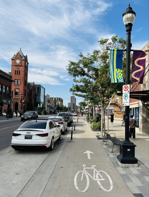 A streetscape featuring a busy sidewalk with parked cars, including a white Audi. A brick clock tower is visible in the background, alongside banners and street lamps. A bicycle lane marked on the pavement runs parallel to the sidewalk, under a partly cloudy sky