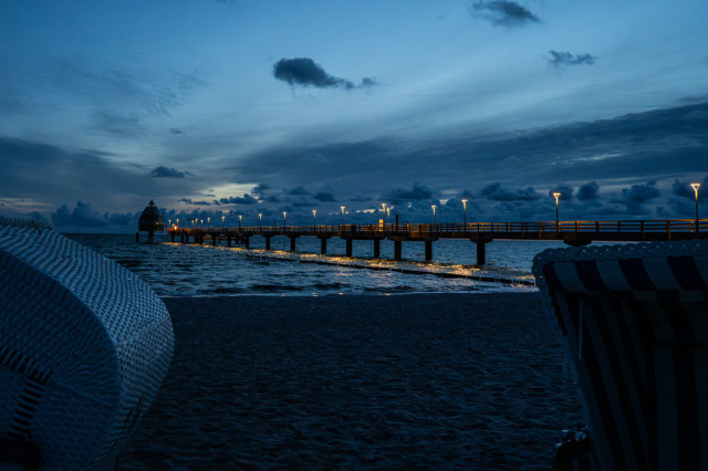 Das Foto zeigt die Seebrücke von Zingst zur blauen Stunde, kurz bevor die Sonne aufgeht. Die Brücke ist beleuchtet, die Lichter spiegeln sich im Wasser der Ostsee. Die Brücke ragt von rechts in das Bild hinein, am Ende lässt sich noch die Tauchgondel erkennen. Der Himmel ist von einigen Wolken durchzogen.
rechts und links unten sind noch Teile von Strandkörben zu sehen. 