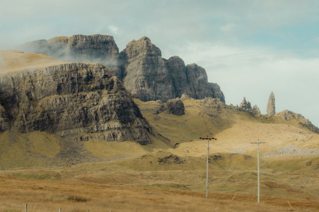 A pale-toned photograph of a rocky mountain and the yellow grassy hills leading up to it, a little mist at its top, against a pale blue sky with whispy clouds. Ther are two telegraph poles in the foreground.