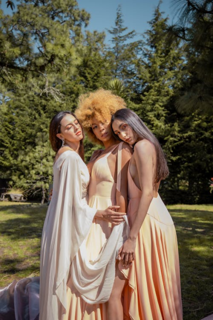 Three fems in long, flowing white and peach silky dresses, grouped together with their arms and heads touching, standing in an open grassy space with some trees behind.