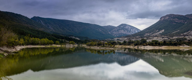 Panorámica de seis fotografías verticales del embalse de Arguis (Huesca). Se puede ver un paisaje nublado, con el agua calmada en la mitad inferior reflejando parte de las montañas de la Sierra de Gratal.