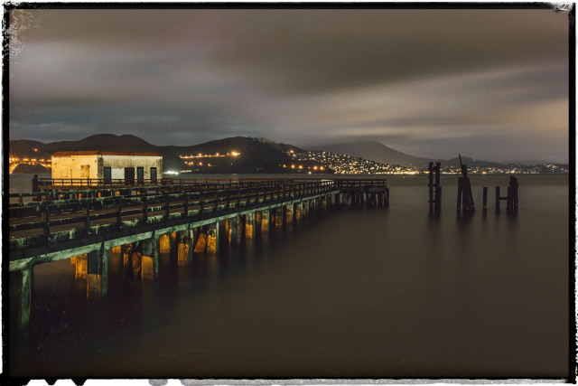 A long wooden fishing pier juts out into the San Francisco Bay. Some wooden pilings also rise up from the water, nearby. Taken at night, the lights from Sausalito are visible in the distance. Also, the pier glows with green and yellow lights, which were barely visible to the naked eye but pop here in this very long exposure.

The cloudy sky and choppy brown bay water are smooth. 153 seconds does that to movement.