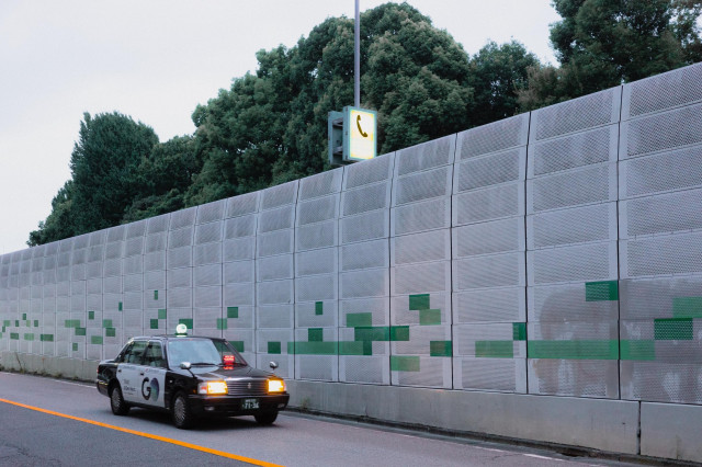 A Japanese taxi drives past a long metal wall with green pixel-like designs. Above the wall are trees, and a yellow illuminated phone booth sign is visible on a pole. The road has an orange line marking.