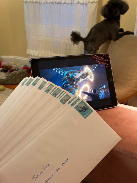 In the foreground a stack of just-completed voter registration letters, stamped, addressed, and ready to be mailed. In the middle ground an iPad playing an Avengers movie. In the background a small curly-haired silver and black dog standing on a chair, looking charmingly over his shoulder, ever hopeful that this somehow involves treats.