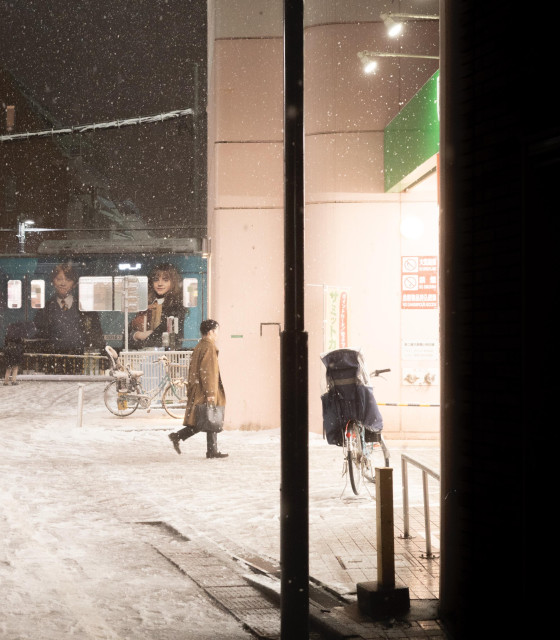 A snowy night scene at a Japanese train station. A person in a brown coat walks through the snow-covered ground. A train is visible in the background with passengers inside. Bicycles are parked near the station entrance. The image captures the soft glow of station lights illuminating falling snowflakes, creating a serene winter atmosphere.