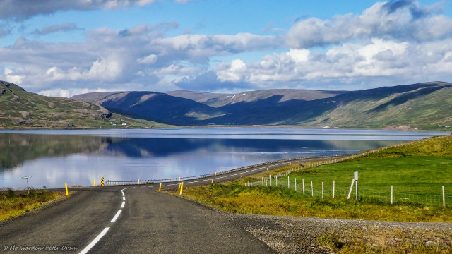 A photo of a landscape, the foreground is a road that we're following with a gravel verge and green meadows. Just beyond a fence is a large body of water and, on the opposite shore, softly rolling hills clothed in summer green. Cloud shadows are scattered across them. The sky has large cyan patches but is half cloudy, with bands of billows in the distance. A few farm buildings are visible on the far shore, and the water is ruffled by feeding birds. It's tranquil and peaceful, and very beautiful.