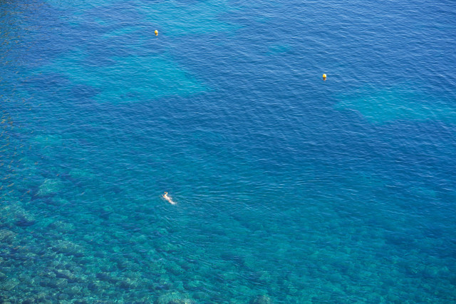 Aerial view of crystal clear turquoise waters. A lone swimmer is visible in the center, surrounded by varying shades of blue sea. The water's clarity reveals a rocky seabed near the shore, transitioning to deeper blue further out. Two yellow buoys float in the distance, marking swimming areas. The scene captures the pristine beauty of a Mediterranean or tropical coastline.