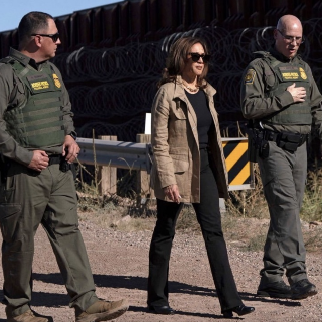 A woman in sunglasses and a beige jacket walks flanked by two uniformed officers wearing green vests and sunglasses. Barbed wire and a fence are visible in the background.