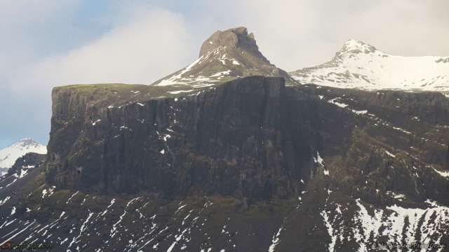 A photo of a mountain, with a cloudy grey sky above. The peaks are two fairly sharp points, one of which is snow-capped. The lower slopes are streaked with ice but the front of the mountain is quite unusual in that it has an odd, slab-like rock standing upright before one of the peaks. This wedge has vertical stripes where the surface is scored, and the top is remarkably flat albeit with a curve. It's quite surreal.