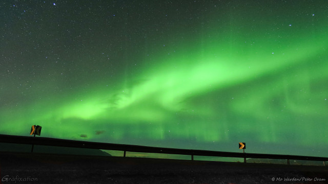 A photo of the night sky with ropes and swirls of aurora, almost in concentric circles. Closer inspection shows that some of the loops have streamers going upward and fading into the night sky. Many stars are visible too. The foreground is a dark stripe which is actually a roadside barrier that we were parked behind. It has yellow chevrons on the top for the traffic on the other side.