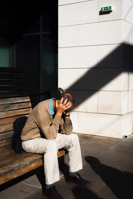 A man sits alone on a wooden bench outside a building, his head in his hands in a gesture of distress or fatigue. He's wearing a beige sweater over a blue checkered shirt and light-colored pants. The scene is dramatically lit with strong sunlight casting sharp shadows across the white tiled wall behind him. An emergency exit sign is visible on the wall above. The contrast between the bright sunlight and deep shadows adds to the emotional weight of the image.