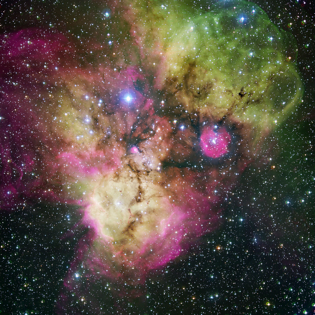 Image of NGC 2467 from the ESO. The image shows a active stellar nursery with dust and gas in pink, green, and yellow shades. The nebula looks like a skull with the left eye a bright blue star and the right eye a pink area of dust/gas. The nebula is amid many smaller white and blue stars. 