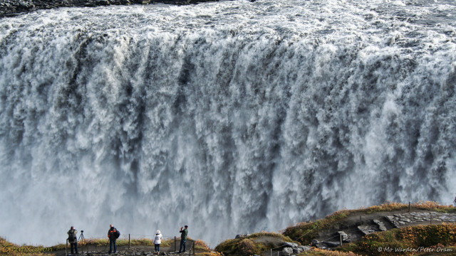 A photo of a massive waterfall, dropping over a quite straight crest line from the top left of the shot to above centre right. The foreground is the canyon's edge, with brown vegetation on either side of a rocky path with steps. Four people are standing in the lower left, all appear to be taking photos. The edge has some open wire fences on low poles. The scene is lit from the right, the far bank is just visible at the top left. The water is aerated and white from the brink to the unseen base. It's awe-inspiring.