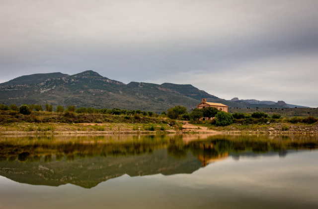 Fotografía donde se ve parte del embalse de Arascués con la ermita de la virgen del Olivar. En la mitad inferior se ven los reflejos y en la superior la ermita con el cielo nublado.