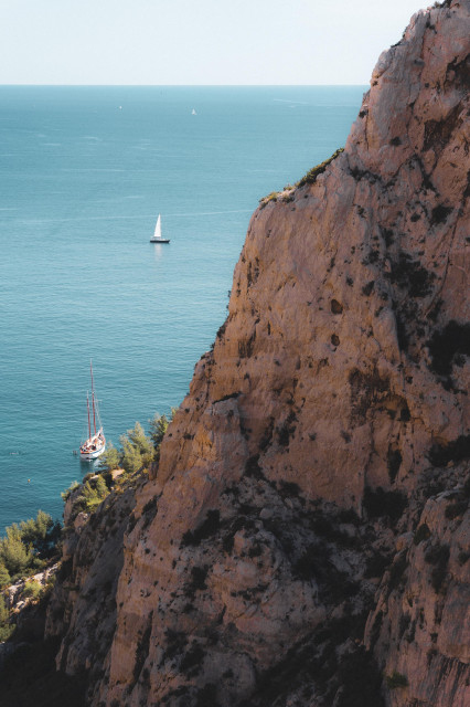 A dramatic coastal scene featuring a towering, rugged cliff face in warm orange hues against a calm turquoise sea. A sailboat is anchored near the base of the cliff, dwarfed by the rock formation. In the distance, other sailboats can be seen on the horizon. The cliff's texture is rough and pockmarked, with sparse vegetation clinging to its surface.