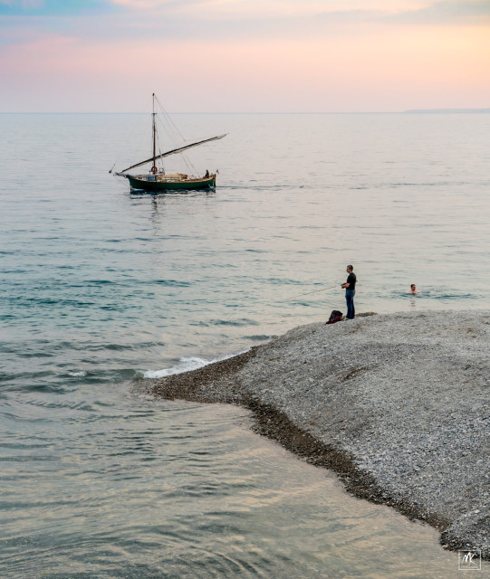 Color photo of a pebbly beach shoreline taken just after sunset with a person fishing from it, another person swimming offshore, and further out a sailboat passing with sails furled. 