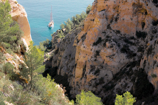 A breathtaking view of a rugged coastal landscape. Massive golden-hued limestone cliffs dominate the foreground, their surfaces pockmarked and textured. Sparse vegetation, including pine trees and shrubs, clings to the rocky terrain. In the distance, a small sailboat is visible on the turquoise waters, nestled between the cliff faces.