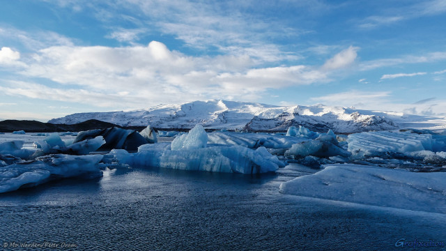 A photo of a large body of blue water with floating icebergs. They are so numerous that they hide most of the water's surface. Some are quite smooth, others are ribbed and most display some darker stripes which are evidence of volcanic ash. The sky is half cloud but quite broken with lots of cyan visible. On the horizon is an imposing mountain, an obtuse triangle covered in ice and snow. The daylight from the left is highlighting the contours of its faces. The entire shot is a study in shades of blue and cyan.