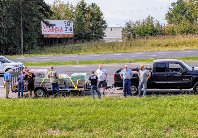 a pickup truck hauling a flatbed trailer with pallets of drinks has pulled off the freeway onto the gravel shoulder, a group of seven men doing various things. four empty lanes behind them with a billboard and trees, etc. it's a semi-rural setting 