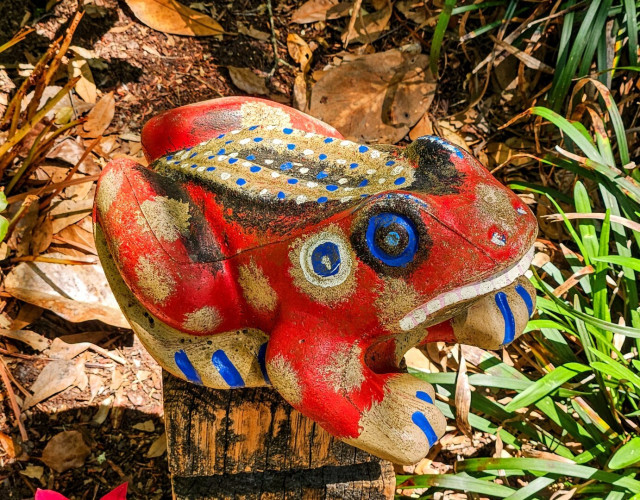 Very near the ground, a large, painted frog decoration sits atop a wooden post. Painted bright red with fanciful spots and colorful circles.