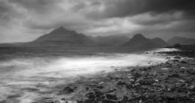 A black and white photo of a stormy sea loch near the village of Elgol, on the Isle of Skye with waves being whipped up looking blurry as it was a long exposure photo. The jagged topped Cuillin mountain range is in the distance under a stormy sky.