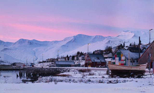 A photo of dawn above a mountain range, with a small town nestled next to a harbour. In the foreground is a snow-covered area where a fishing boat sits on a launching dolly. Behind it is a row of houses and a line of lit street-lights disappearing around a corner. The mountains beyond are completely snow-capped and are in shades of blue and cyan in the increasing light. The sky above is streaked with beautiful pastel shades of cyan, mauve and pink.