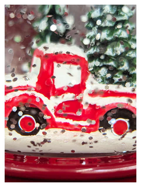 close-up view of a snow globe on a store shelf, glitter shaken for effect. the base is shiny crimson plastic, the scene inside is a vintage red pickup truck with two large evergreens behind it. snow dusts or covers everything.