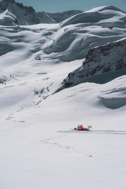 A vast, snow-covered mountain landscape with jagged peaks and smooth, undulating snow fields. In the foreground, a small red snowcat vehicle traverses the expansive white terrain, leaving a thin trail behind it. The scale of the mountain dwarfs the vehicle, emphasizing the grandeur and isolation of the wintry environment.