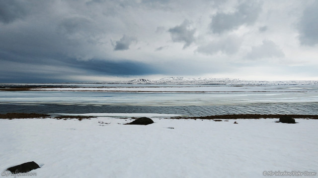 A photo of a frozen landscape in shades of cyan. The foreground is an untrammelled snow field with black rocks poking through it. Beyond is a narrow strip of open water, the rest of the surface is frozen. After that is a black sand bar, covered in ice and snow except for the edges and then another stretch of thickly iced water. In the distance is a shoreline with a mountain range visible beyond. The sky is full of heavy cloud, some very dark.