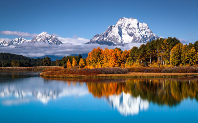 A wide calm river reflecting trees and mountains and blue sky beyond. Leaves on the trees are changing to fall colors. High steep mountains are capped with snow. Location is Oxbow Bend, Jackson Hole, Wyoming, at sunrise.