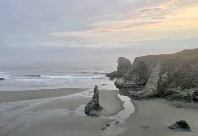 A serene beach scene at dusk, featuring gentle ocean waves, misty skies, and rocky coastal formations. A tall rock spire stands prominently in the foreground, with a smooth sandy beach stretching out beside it.