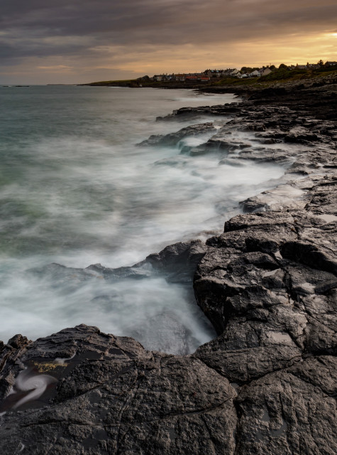 Waves crashing on the black rocls of the Whin Sill with Craster in the background.