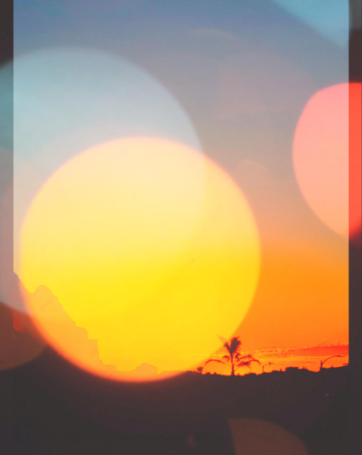 An abstract photo of a sunset. In the far distance we see a lone palm tree standing tall above houses and street lights across a darkened San Francisco skyline—all rendered in silhouette against a brilliant red-orange sun which is setting below a blueish-grey early evening sky. Huge multi-color bokeh bubbles of yellow, blue, orange, and red float across the scene giving an ethereal, dream-like style to the image. The ends of a very warm day, playing beautiful tricks with my eyes …as the City finally cools enough to enjoy a beer on the porch with the neighbors!