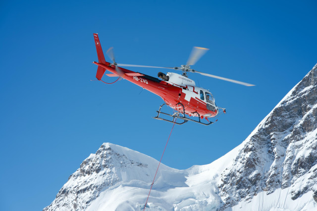 A red and white Swiss rescue helicopter flies against a bright blue sky, with snow-covered mountain peaks in the background. The helicopter, marked with the Swiss flag and registration number HB-ZID, is in mid-flight with its rotors blurred from motion. A thin red cable or rope can be seen extending from the bottom of the aircraft. The rugged, snow-capped mountains provide a dramatic backdrop, emphasizing the helicopter's role in alpine rescue operations.